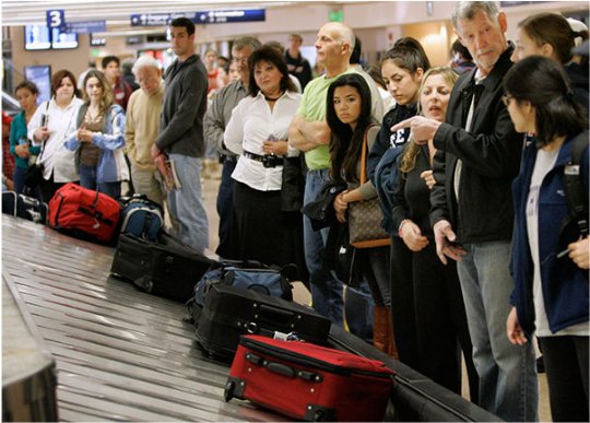 Image of an airport baggage claim carousel with people waiting for their bags.